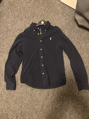 Mörkblå skjorta från Polo Ralph Lauren - Klassisk mörkblå skjorta från Polo Ralph Lauren med button-down krage och broderad logga på bröstet. Skjortan har långa ärmar och vita knappar framtill samt vid manschetterna. Perfekt till jeans eller chinos. Säljes då den inte passade mig. Inte använd.