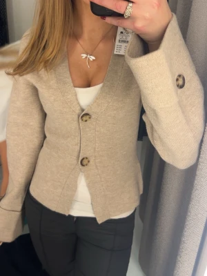 Beige kofta med stora knappar - Trendig beige kofta med två stora, mönstrade knappar framtill och vida ärmar. Koftan har en djup V-ringning och är figursydd för en snygg siluett. Perfekt att styla över ett linne eller t-shirt. Materialet ser ut att vara mjukt och stickat.