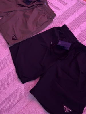 Svarta shorts från Prada - 350kr per shorts, 650kr för bundle. Svarta shorts från Prada med elastisk midja och snörning. Klassisk design med diskret Prada-logga på benet. Materialet är syntet och ger en lätt känsla, perfekt för varma dagar. Passformen är normal och stilen är enkel men exklusiv.