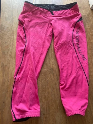 Rosa Better Bodies leggings - Snygga korta rosa leggings från Better Bodies med svarta detaljer och logga längs benet. De har en tight passform och är perfekta för träning eller chill. Materialet är stretchigt och bekvämt, och det finns svarta inslag vid linningen och längs benen.