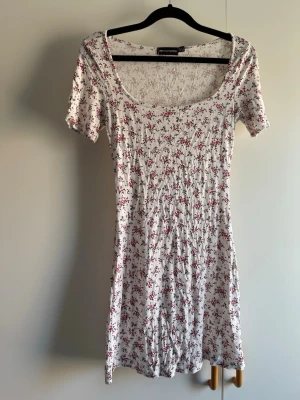 Vit blommig klänning från PrettyLittleThing - Söt vit kortklänning från PrettyLittleThing med små rosa och röda blommor. Klänningen har korta ärmar, rundad urringning och är tillverkad i ett mjukt bomullstyg. Perfekt för varma dagar och en avslappnad look.
