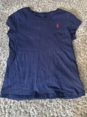 Marinblå t-shirt från Ralph Lauren - Klassisk marinblå t-shirt från Ralph Lauren med röd broderad logga på bröstet. T-shirten har rund halsringning och korta ärmar. Tillverkad i mjuk bomull som känns skön mot huden. Perfekt basplagg till garderoben.