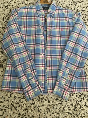 Rutig skjorta från Polo Ralph Lauren - Säljer en klassisk rutig skjorta från Polo Ralph Lauren i blått, vitt, rött och grönt. Skjortan har långa ärmar, knappar framtill och den ikoniska röda loggan broderad på bröstet. Perfekt för dig som gillar preppy stil och vill ha något snyggt till garderoben.