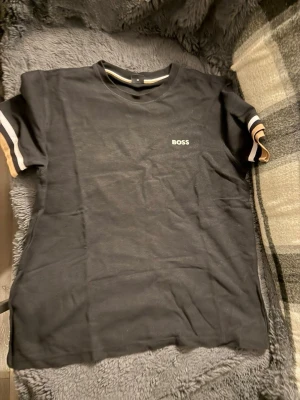 Svart BOSS t-shirt med randiga ärmar - Svart t-shirt från BOSS med korta ärmar och diskret logga på bröstet. Ärmarna har snygga ränder i vitt och beige som ger en sportig touch. Tillverkad i mjuk bomull och har en klassisk passform.