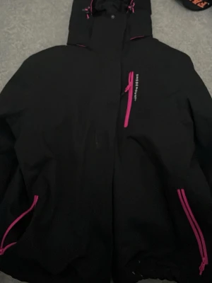 Svart skidjacka med rosa detaljer - Snygg svart skidjacka från swedemount med rosa dragkedjor och detaljer. Jackan har huva, flera fickor med dragkedja och är fodrad på insidan med rosa partier. Perfekt för en kall skidtur om man ska ut i år 