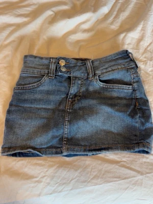 Blå jeanskjol med dubbla knappar - Snygg blå jeanskjol i klassisk modell med dubbla knappar framtill, fem fickor och bälteshällor. 