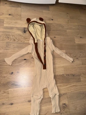 Beige onesie med öron och knappar - Supersöt beige ribbad onesie med huva som har små bruna öron. Knappar framtill med bruna detaljer och mjukt tyg på insidan av huvan. Perfekt för mysiga hemmadagar eller som loungewear för de minsta.