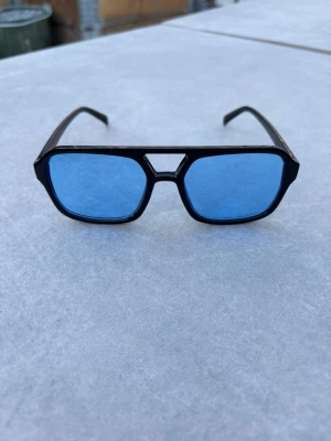 Svarta solglasögon med blåa glas - Coola solglasögon med svart ram och blåa, fyrkantiga glas. Ramen har en unik detalj vid näsryggen och glasen ger en snygg kontrast. Perfekt accessoar för dig som vill sticka ut med en retrovibe.