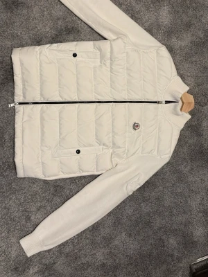 Vit cardigan från Moncler - Snygg vit pufferjacka från Moncler med stickade ärmar och quiltad front. Jackan har dragkedja, två fickor med knappstängning och Moncler-logga på bröstet. Perfekt för dig som vill ha en stilren och modern look.