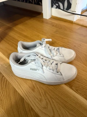 Vita Puma sneakers med grå detaljer - Fräscha vita sneakers från Puma med klassisk låg profil och grå detaljer. Skorna har vita skosnören, diskret Puma-logga på sidan och är tillverkade i syntetmaterial med mjuk vadderad insida. Perfekta för en clean och sportig look. Storlek 37