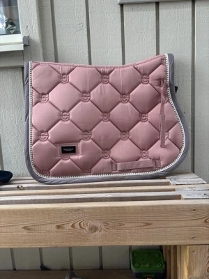Rosa schabrak från Equestrian Stockholm - Säljer ett snyggt schabrak i ljusrosa från Equestrian Stockholm. Schabraket har quiltat mönster med små loggor, grå kant och dekorativ passpoal. Perfekt för dig som vill ha stil på ridturen. Använt en gång, så i skit bra skick. Pris diskuterbart💞