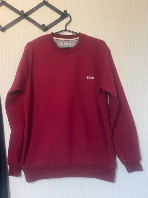 Vinröd sweatshirt från BOSS - Snygg vinröd sweatshirt från BOSS med rund halsringning och diskret BOSS-logga broderad i vitt på bröstet. Tröjan har ribbade muddar vid ärmslut och nederkant. Perfekt för en clean och stilren look.