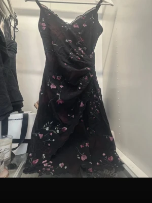 Svart blommig klänning - Säljer en svart kortklänning med tunna axelband.  Klänningen har ett blommigt mönster i rosa och grönt samt volangkant nedtill. Perfekt för dig som gillar romantiska detaljer och en feminin look.