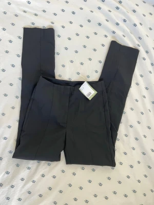 Svarta kostymbyxor från H&M - Snygga svarta kostymbyxor från H&M med klassisk rak passform och pressveck längs benen. Byxorna har hög midja och dold knäppning framtill. Perfekta för en stilren look och enkla att matcha med olika överdelar.