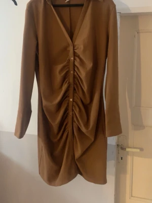 Brun omlottklänning från H&M - Snygg brun kortklänning från H&M med långa ärmar och knappar framtill. Klänningen har en omlott-effekt och rynkade detaljer längs hela framsidan som ger en cool och trendig look. Perfekt för dig som gillar stilrena och moderna plagg.