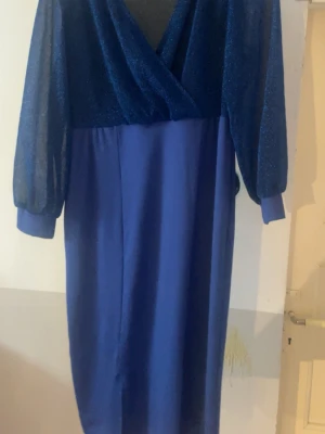 Blå långklänning med glitterärmar - Snygg blå långklänning med glittriga, transparenta långärmar och omlott-v-ringning. Klänningen har en elegant och modern look, perfekt för dig som vill sticka ut. Materialet känns mjukt och bekvämt, och ärmarna ger en extra touch av glamour.