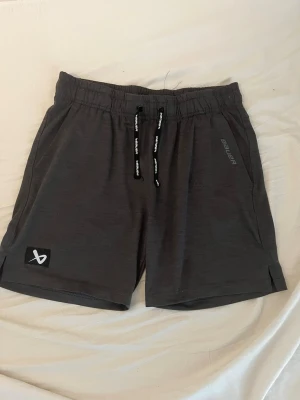 Mörkgrå shorts från Bauer - Snygga mörkgrå shorts från Bauer med elastisk midja och svarta snören med logga. Shortsen har en diskret Bauer-logga på benet och en liten svart patch med vit symbol. Perfekta för träning eller chill.
