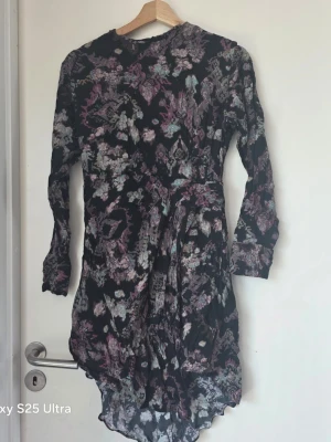 Svart blommig långärmad klänning - Svart klänning med abstrakt blommigt mönster i lila, turkos och grå toner. Långärmad modell med asymmetrisk nederkant och rund halsringning. Perfekt för dig som gillar unika prints och en bekväm passform.