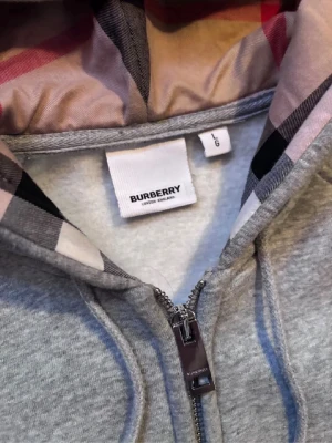 Grå zip hoodie från Burberry - Snygg grå zip hoodie från Burberry med klassiskt rutigt foder i huvan och dragsko. Tillverkad i mjukt material och har metall-detaljer på snörena samt dragkedja framtill. Perfekt för en chill och stilren look.