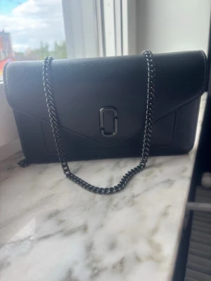 Svart kuvertväska från Marc Jacobs - Snygg svart kuvertväska från Marc Jacobs i strukturerat läder med kedjerem i metall. Väskan har ett stilrent och minimalistiskt utseende med logotypdetalj på framsidan. Perfekt att bära crossbody eller på axeln för en trendig look.