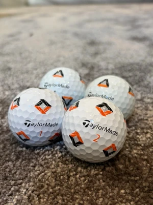 TaylorMade vita golfbollar med tryck - Fyra vita golfbollar från TaylorMade med svart och orange tryck samt numrering. Bollarna har klassisk dimple-design och är perfekta för golfspelare som vill ha stilrena och synliga bollar på banan. Skriv privat för pris o hur många du ska ha