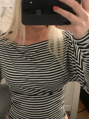 Randig långärmad topp med droppad axel - Snygg långärmad topp med svarta och vita ränder och droppad axel. Modellen är figurnära med bred halsringning och extra långa ärmar. Perfekt för en trendig och avslappnad look.