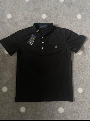Svart pikétröja från Polo Ralph Lauren - Klassisk svart pikétröja från Polo Ralph Lauren med vit broderad logga på bröstet. Tröjan har korta ärmar, krage och knäppning med tre vita knappar. Tillverkad i bomull för en bekväm känsla och snygg passform.