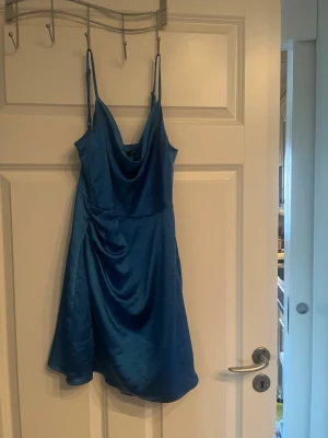 Blå satin klänning med tunna axelband - Snygg blå kort klänning i satin med tunna axelband och draperad urringning. Klänningen har en glansig finish och en lätt utställd passform som ger en elegant siluett. Perfekt för dig som vill sticka ut med en färgstark look.