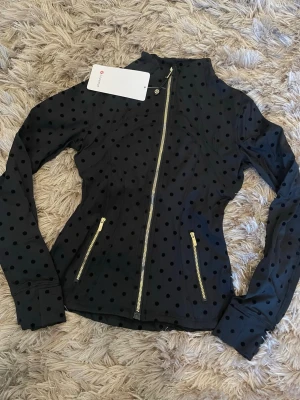  Lululemon nya modellen st 4 - Snygg svart jacka från Lululemon med prickigt mönster och guldiga dragkedjor. Jackan har figurnära passform, hög krage och två fickor framtill. Perfekt för dig som gillar sportig och trendig stil.