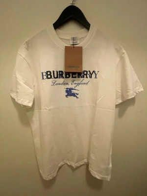 Vit Burberry t-shirt med blå logga - Vit t-shirt från Burberry med blå och svart logotyp och texten 'London, England' framtill. Klassisk rund halsringning och korta ärmar. Tillverkad i mjuk bomull, perfekt för en stilren och trendig look.