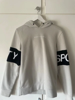 Ljusgrå hoodie med tryck - Ljusgrå hoodie med svart och vitt tryck på ärmarna där det står 'SPORTY'. På bröstet finns koordinaterna 55.9427°N 9.1288°E tryckta i vitt. Hoodien har dragsko i huvan och ribbade muddar. Perfekt för en avslappnad streetwear-look.