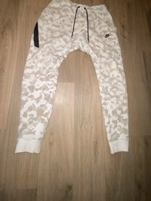 Nike camo byxor med dragsko - Säljer ett par snygga byxor från Nike med beige och vit camomönster. Byxorna har resår i midjan med svart dragsko, muddar vid bensluten och en svart detalj vid fickan. Perfekta för en avslappnad och sportig stil.