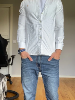 Vit skjorta med knappar från Ralph Lauren - Klassisk vit skjorta från Ralph Lauren med knappar framtill och diskret broderad logga på bröstet. Skjortan har lång ärm och slimmad passform, perfekt till jeans eller chinos. Tillverkad i bomull för en skön känsla och stilren look.