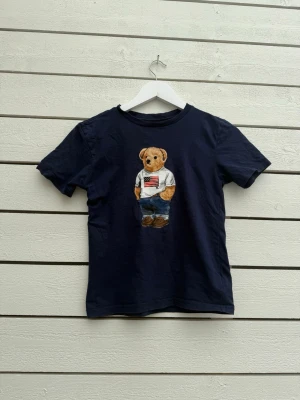 Mörkblå t-shirt med björntryck från Ralph Lauren - Mörkblå t-shirt från Ralph Lauren med ikoniskt björntryck på bröstet. Björnen har på sig jeans och en vit t-shirt med amerikansk flagga. Klassisk rund hals och korta ärmar. Perfekt för en avslappnad och cool stil.