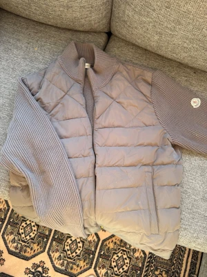 Grå Moncler Cardigan - Snygg grå pufferjacka från Moncler med stickade ärmar och hög krage. Jackan har två fickor framtill och dragkedja. Moncler-logga på ena ärmen. Perfekt för kalla dagar eller sena kvällar, en riktigt trendig look. Jackan är i princip oanvänd! Säljer den åt min kusin men har ni några frågor är de bara att höra av er. Priset kan diskuteras vid snabb affär!!
