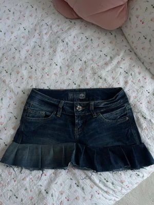 Mörkblå jeanskjol med volangkant - Trendig mörkblå jeanskjol med volangkant nertill och råa kanter. Klassisk femficksmodell med knapp och dragkedja framtill. Perfekt för dig som gillar en edgy och cool stil.