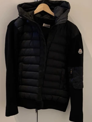 Moncler cardigan - Snygg svart pufferjacka från Moncler med quiltad front och huva. Jackan har stickade ärmar och Moncler-logga på vänster ärm. Dragkedja framtill och praktiska fickor. Perfekt för kalla vinterdagar och riktigt trendig look.