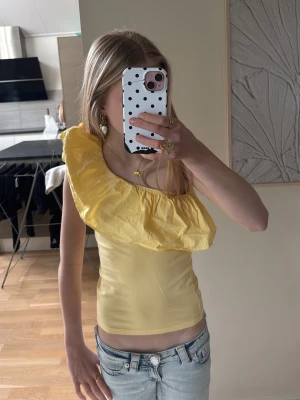 Gul oneshoulder topp med volang - Lappen är klippt av, vet inte riktigt storleken men passar bra på mig, 161 cmSnygg gul oneshoulder topp med stor volangdetalj längs halsringningen. Toppen är figurnära och har en fräsch, somrig vibe. Perfekt för dig som vill sticka ut lite extra med en unik design.