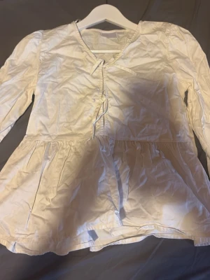 Vit blus med knyt från Gina Tricot - Söt vit blus från Gina Tricot med lång ärm och knytning framtill. Blusen har en lätt v-ringning och volang nedtill som ger en feminin touch. Tillverkad i ett tunt och mjukt tyg, perfekt för en avslappnad och trendig look.