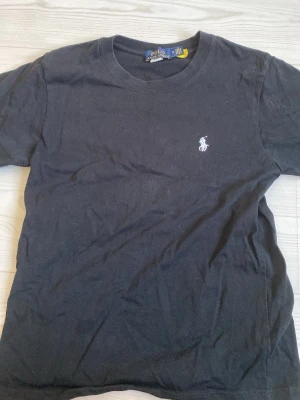 Svart t-shirt från Polo Ralph Lauren - Svart t-shirt från Polo Ralph Lauren med klassisk passform och broderad vit logga på bröstet. Tillverkad i mjuk bomull och har rund halsringning samt korta ärmar. Perfekt för en clean och stilren look.