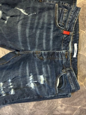 Blå distressed jeans från 2Y Premium - Säljer ett par blå jeans från 2Y Premium med slitningar och distressed detaljer på framsidan. Jeansen har klassisk femficksdesign, orangea sömmar och en cool röd tag vid linningen. Materialet är denim och passformen är rak.