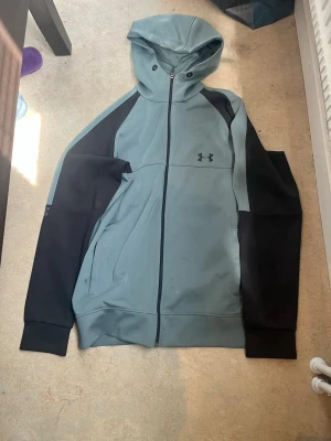 Under Armour hoodie Grön (Sälsynt)  - Snygg hoodie från Under Armour i grått med svarta ärmar och detaljer. Dragkedja framtill, två fickor och klassisk huva. Perfekt för en sportig och avslappnad stil. Materialet är mjukt och stretchigt för extra komfort. De är storlek M men sitter mer som S och XS. Tipen är lite trög men den funkar fortfarande 
