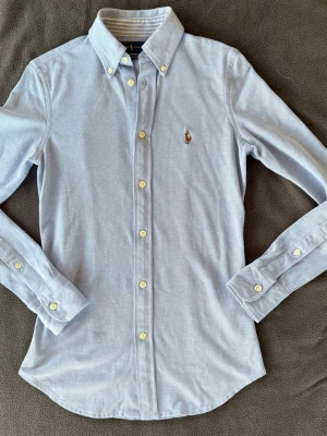 Ljusblå skjorta från Ralph Lauren - Klassisk ljusblå skjorta från Ralph Lauren med broderad logga på bröstet. Skjortan har knappar hela vägen, button-down krage och långa ärmar. Tillverkad i mjuk bomull och har en tidlös, normal passform. Modellen är knot Oxford.