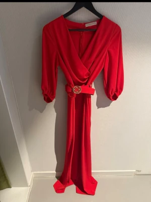 Röd byxdress från Rinascimento - Röd byxdress från Rinascimento med omlott-ringning, långa puffärmar och avtagbart bälte med dekorativt spänne. Tillverkad i ett mjukt, följsamt tyg som ger en snygg siluett. Dragkedja bak. Står XS, men passar S. 