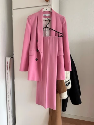 Rosa kostym från Zara - Säljer en snygg rosa kostym från Zara med dubbelknäppt kavaj och matchande byxor. Kavajen har svarta knappar, två fickor och en klassisk krage. Perfekt för dig som vill sticka ut med färg. Materialet känns mjukt och har en lätt struktur.