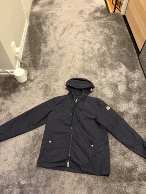 Mörkblå vindjacka från Moncler - Snygg mörkblå vindjacka från Moncler med huva och dragkedja framtill. Jackan har två fickor med tryckknappar och en vit-röd rand på huvan. Moncler-logga på vänster ärm. Perfekt för blåsiga dagar.