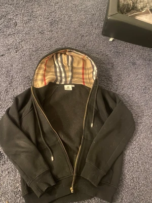Svart hoodie från Burberry med dragkedja - Svart hoodie från Burberry med klassiskt rutigt foder i huvan och guldfärgad dragkedja. Hoodien har långa ärmar, ribbade muddar och en stilren look. Perfekt för dig som vill ha en lyxig streetstyle-vibe. Hör av er vid frågor eller funderingar 