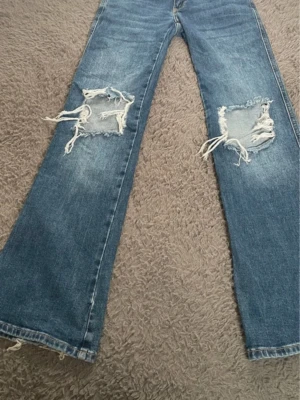Blå slitna jeans från 157 - Ett par blå jeans från 157 med raka ben och stora slitningar över båda knäna. Jeansen har klassisk femficksdesign, dubbla knappar i midjan och tydliga slitdetaljer för en trendig look.