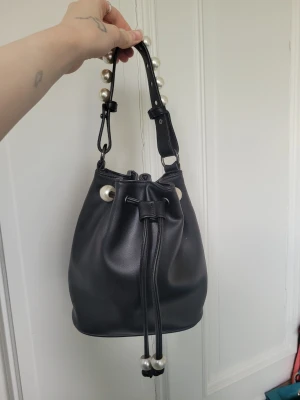 Svart bucket bag med pärldetaljer - Trendig svart bucket bag i slätt konstläder med dragskostängning och stora pärldetaljer på handtaget. Så fin och får plats med mer än vad man tror! Toppen skick!🩷
