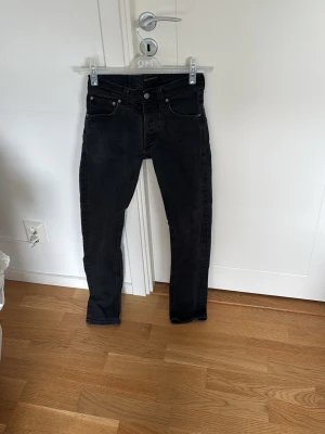 Svarta slim jeans från nudie - Säljer ett par svarta slim jeans från nudie med klassisk femficksdesign och knappgylf. Jeansen har en tight passform och är tillverkade i stretchigt denimtyg för extra komfort. Perfekta för dig som gillar en stilren och enkel look.